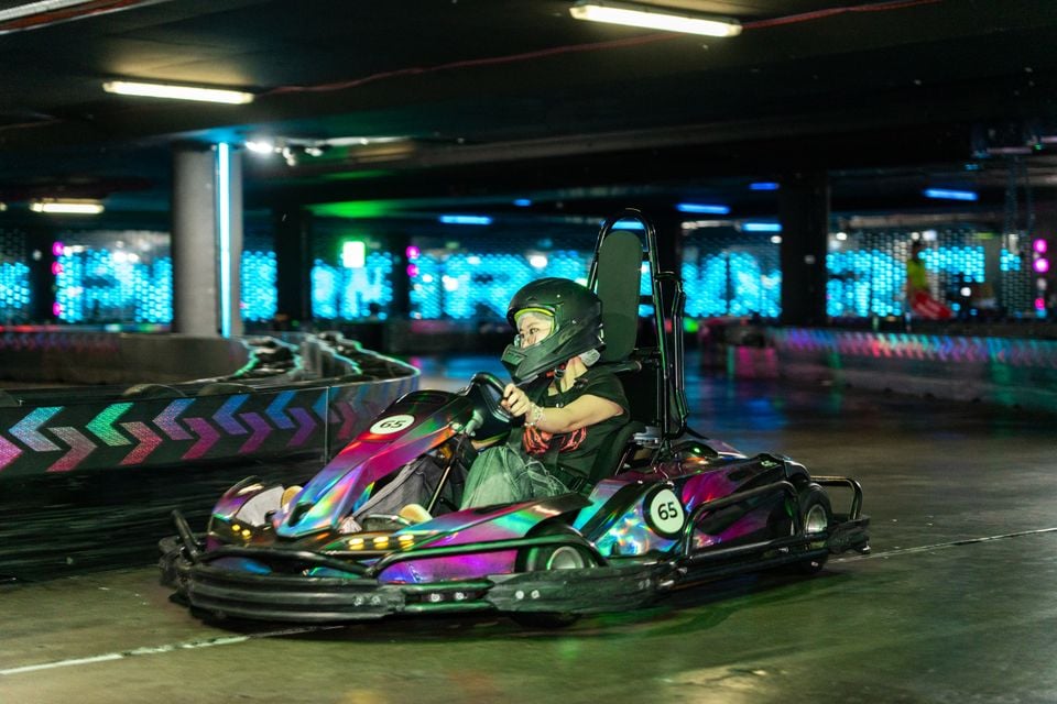 A person wearing a helmet drives a holographic go-kart numbered 65 around an indoor karting track with colourful neon lighting.