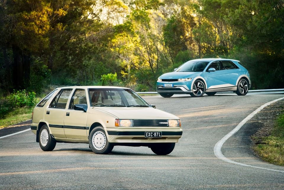 A classic cream Hyundai Excel hatchback parked in the foreground alongside a blue Hyundai Ioniq 5 on a winding road surrounded by Australian bushland
