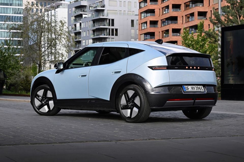 A light blue Hyundai Ioniq electric SUV parked on a cobblestone street, seen from the rear quarter with city apartment buildings in the background.