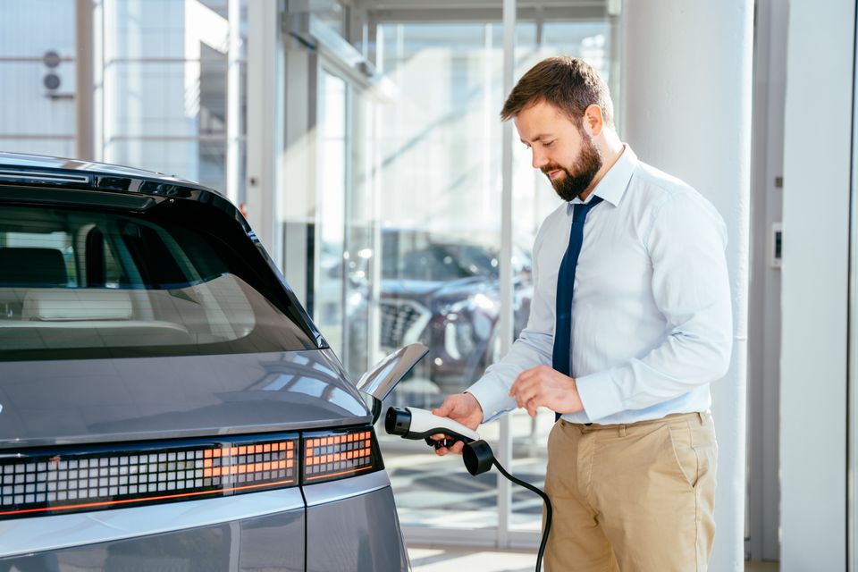 A man in a shirt and tie holds an EV charging connector at the rear charge port of a grey Hyundai Ioniq 5 inside a dealership showroom.