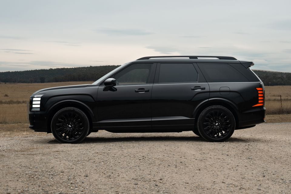 A black Hyundai Palisade large SUV seen from the side, parked on a gravel road with an open rural landscape behind it