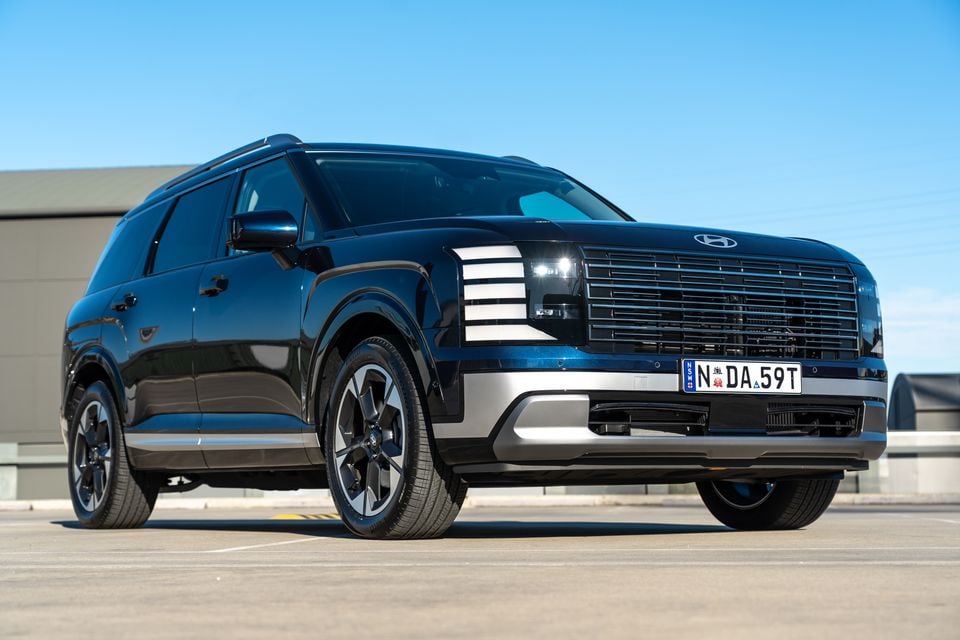 A dark blue Hyundai Palisade large SUV seen from the front-side angle in a car park under a clear blue sky