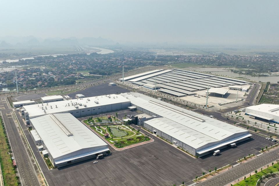 Aerial view of a large automotive manufacturing facility with multiple warehouse buildings, landscaped gardens, and surrounding roads, with a town and river visible in the background.
