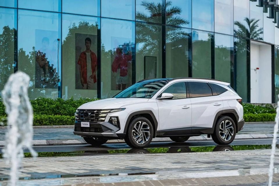 A white Hyundai Tucson SUV parked outside a modern glass building with water fountains in the foreground, seen from the front-side angle.