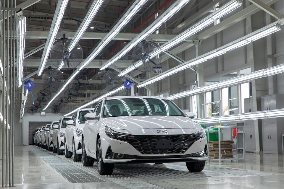 A row of white Hyundai Elantra sedans lined up on a production line inside a brightly lit factory.