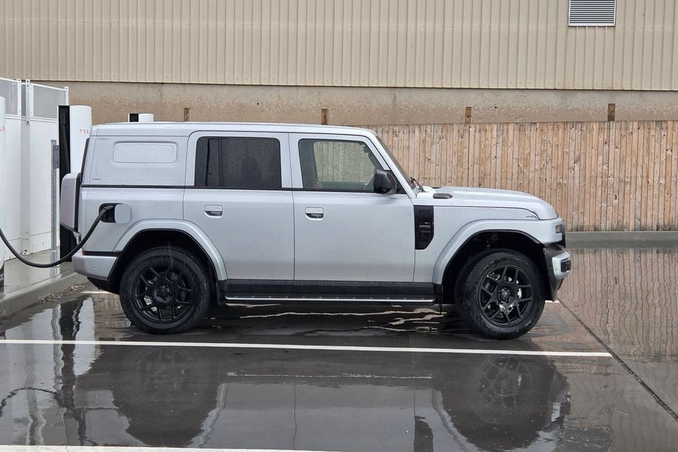 A silver electric SUV plugged into a Tesla Supercharger at a wet carpark, seen from the side