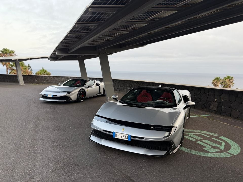Two silver Ferrari 296 GTS spider supercars parked under a solar panel canopy with ocean views in the background, red interior visible