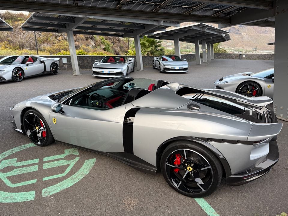 Multiple silver Ferrari sports cars parked under a covered area with solar panels, with a mountainous landscape in the background — the closest car is a silver open-top Ferrari spider seen from the rear side