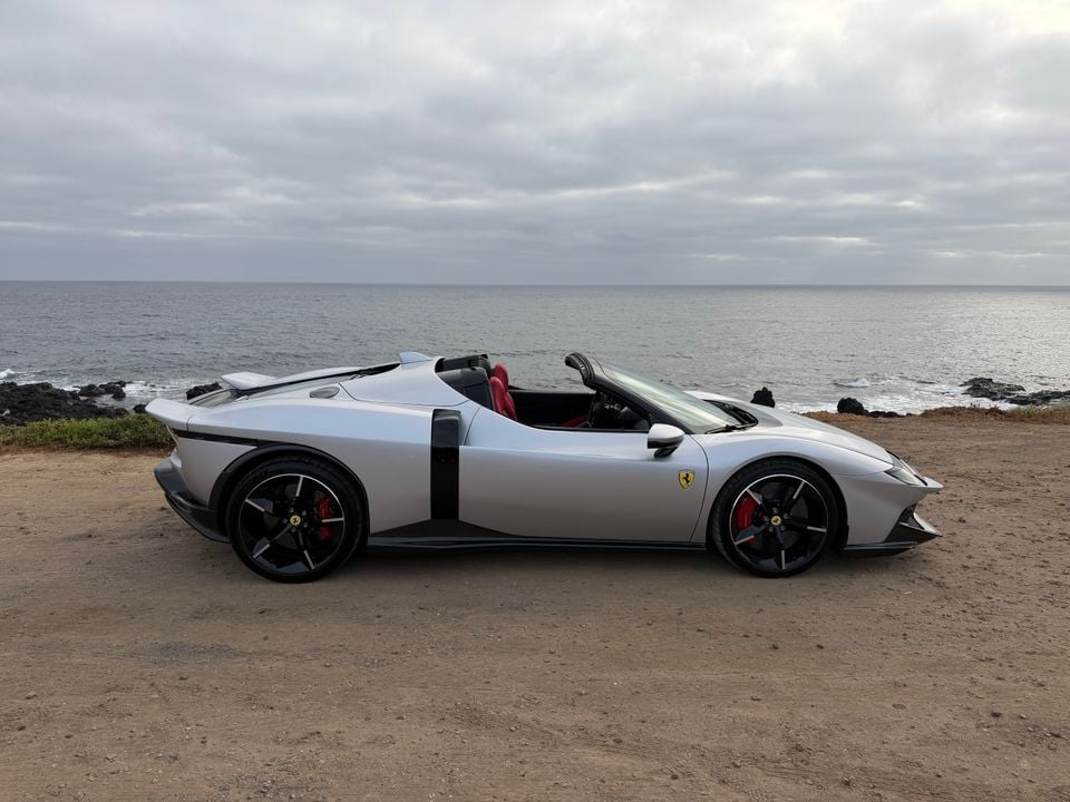 A silver Ferrari open-top sports car seen from the side, parked near a rocky coastline under an overcast sky