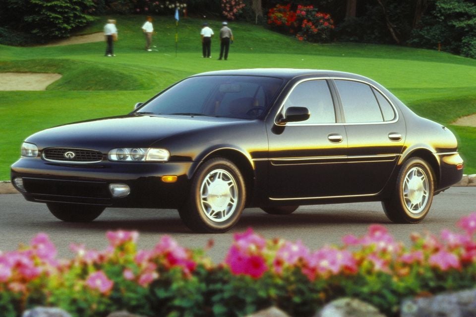A black Infiniti J30 sedan seen from the front-side angle, parked near a golf course with flowers in the foreground.