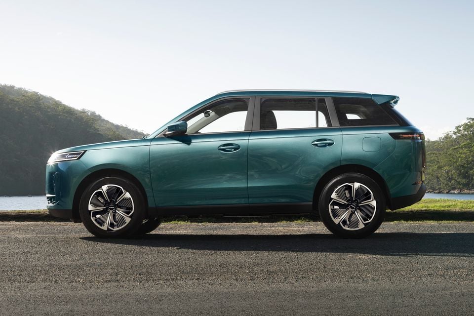 A teal Jaecoo J5 hybrid SUV seen from the side, parked near a waterway with trees in the background