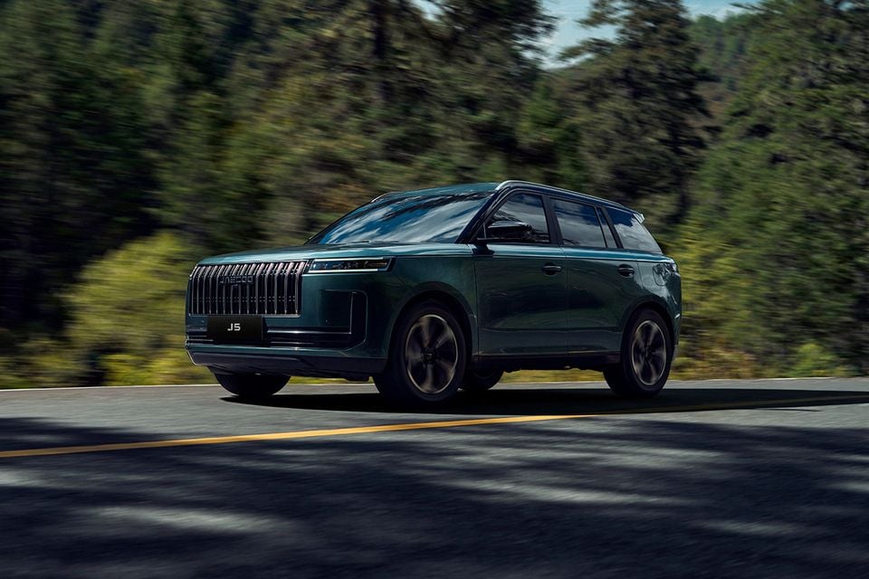 A green Jaecoo J5 SUV driving along a road with a forest backdrop, seen from the front-side