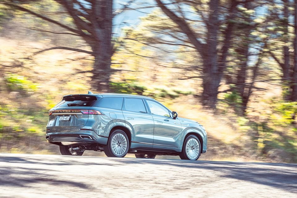 A grey Jaecoo J8 SUV seen from the rear three-quarter angle, driving along a tree-lined road