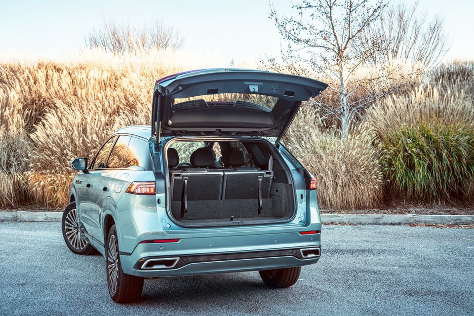 A light blue Jaecoo J8 SUV seen from the rear with the tailgate open, revealing the boot space, parked outdoors near golden grasses.