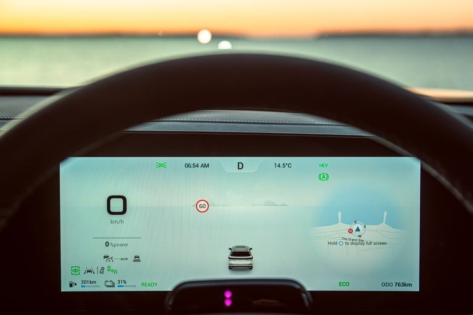 Close-up of a Jaecoo J8 digital instrument cluster showing speed, HEV hybrid mode, eco driving mode, and a navigation map, with a coastal sunset visible through the windscreen