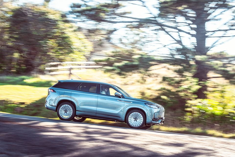A light blue Jaecoo J8 SUV driving along a tree-lined road, seen from the side
