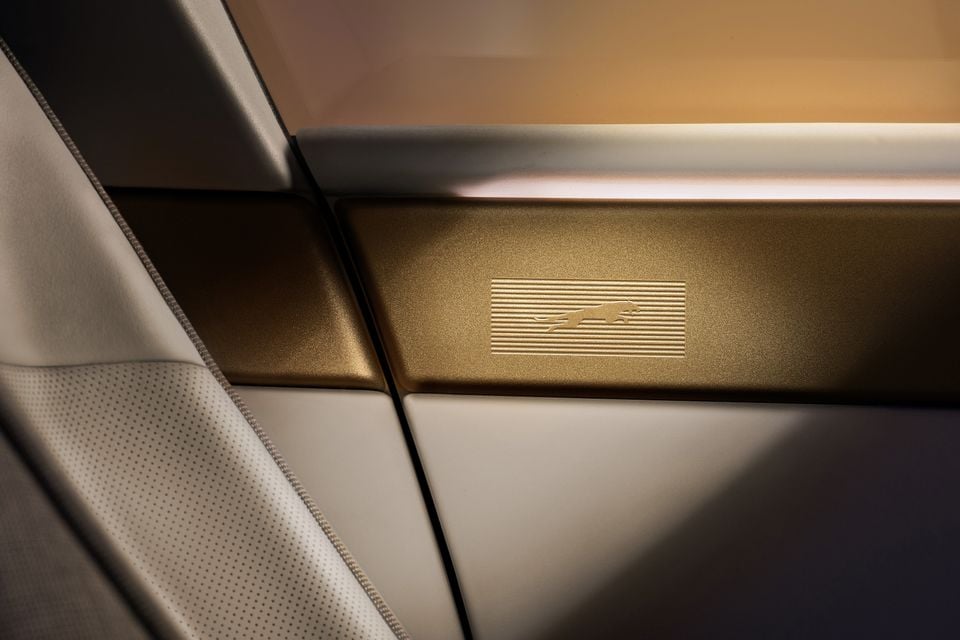 Close-up of a Jaguar Type 00 interior door panel featuring a gold-toned trim strip with an embossed Jaguar leaping cat logo, alongside cream leather upholstery