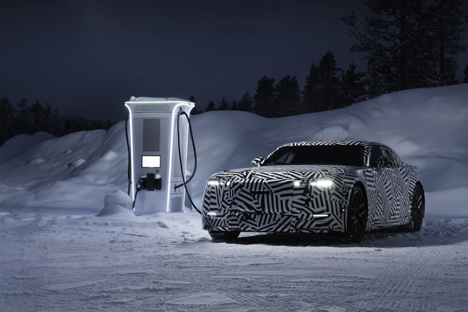A camouflaged Jaguar Type 00 electric sedan prototype parked next to an illuminated fast-charging station in a snowy winter landscape at night, with headlights on
