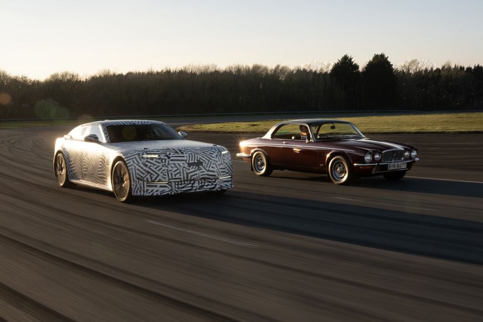 A camouflaged Jaguar Type 00 prototype sedan driving alongside a classic dark red Jaguar XJ coupe on a test track at dusk.