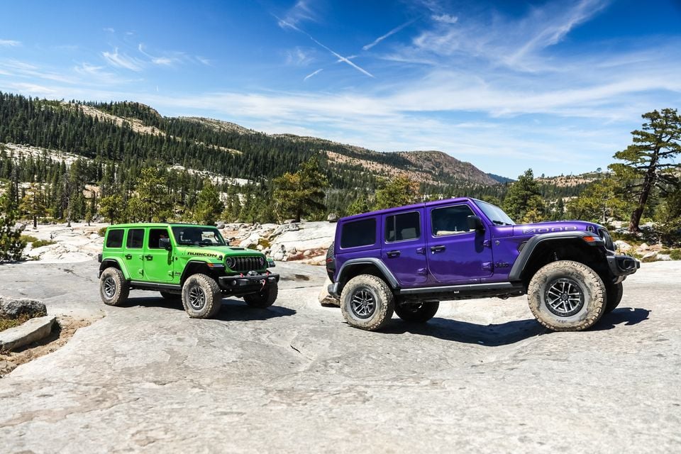 A green Jeep Wrangler Rubicon and a purple Jeep Wrangler Moab parked on granite rock on the Rubicon Trail, with pine-covered hills behind