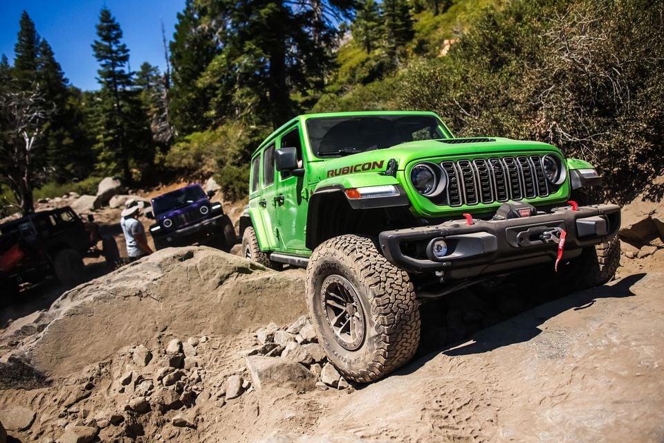 A green Jeep Wrangler Rubicon 392 crawling over rocks on the Rubicon Trail, with other Jeeps and a spotter behind among pine trees