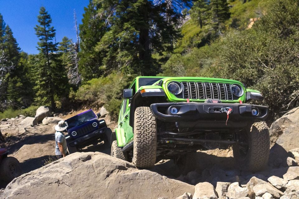 A green Jeep Wrangler 392 crawling over boulders on the Rubicon Trail, with a purple Wrangler and a spotter following behind among pine trees