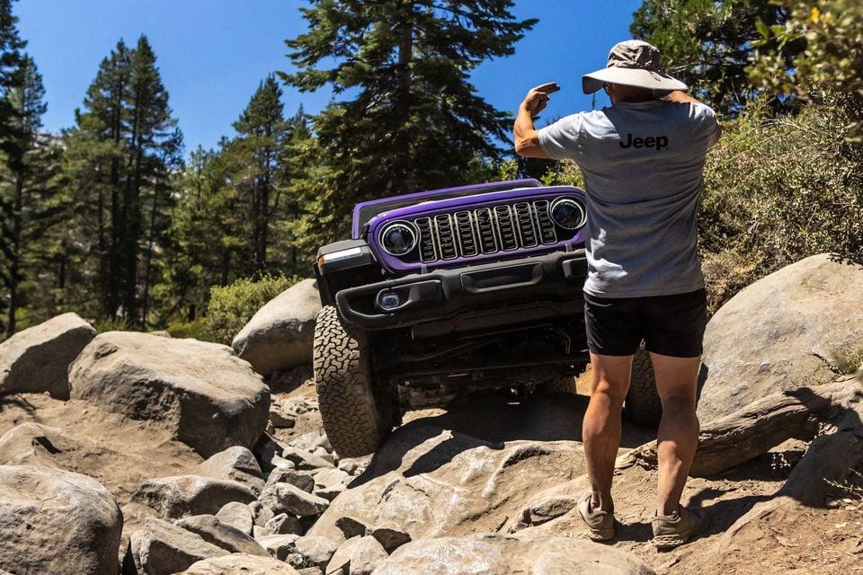 A purple Jeep Wrangler 392 crawling over boulders on the Rubicon Trail as a spotter in a Jeep shirt guides the driver