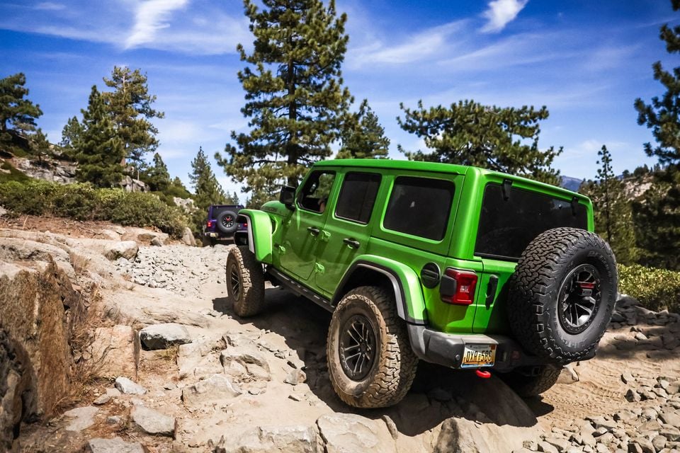A green Jeep Wrangler Rubicon 392 seen from the rear crawling over rocks on the Rubicon Trail, with a purple Wrangler ahead among pine trees