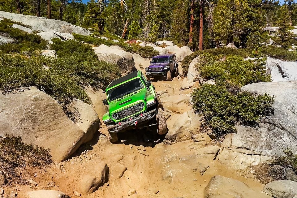 A green and a purple Jeep Wrangler 392 crawling over granite boulders on the Rubicon Trail, seen from the front among pine trees