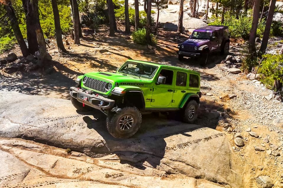 A green Jeep Wrangler Rubicon 392 crawling over granite rock on the Rubicon Trail, followed by a purple Jeep Wrangler among pine trees