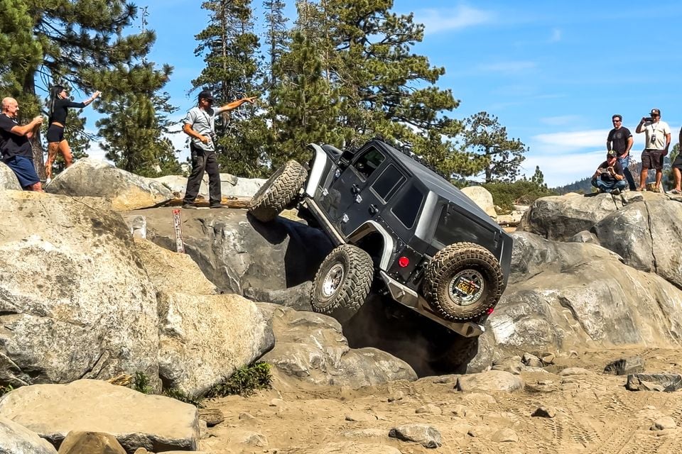 A dark grey two-door Jeep Wrangler climbing a steep granite boulder on the Rubicon Trail, seen from the rear as spectators watch and take photos