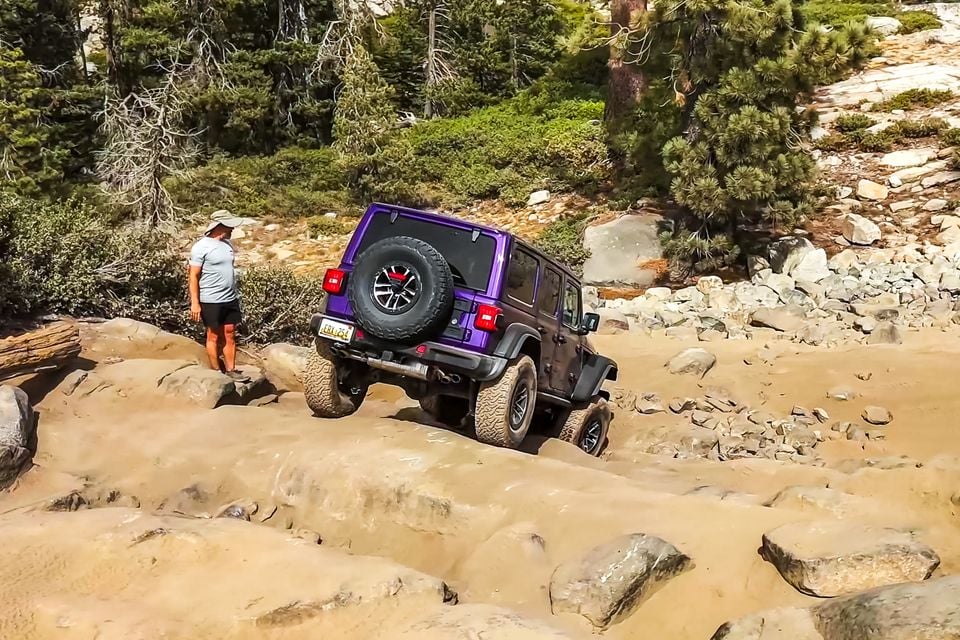 A purple Jeep Wrangler 392 four-door climbing over granite rocks on the Rubicon Trail, seen from the rear, as a man watches beside the track