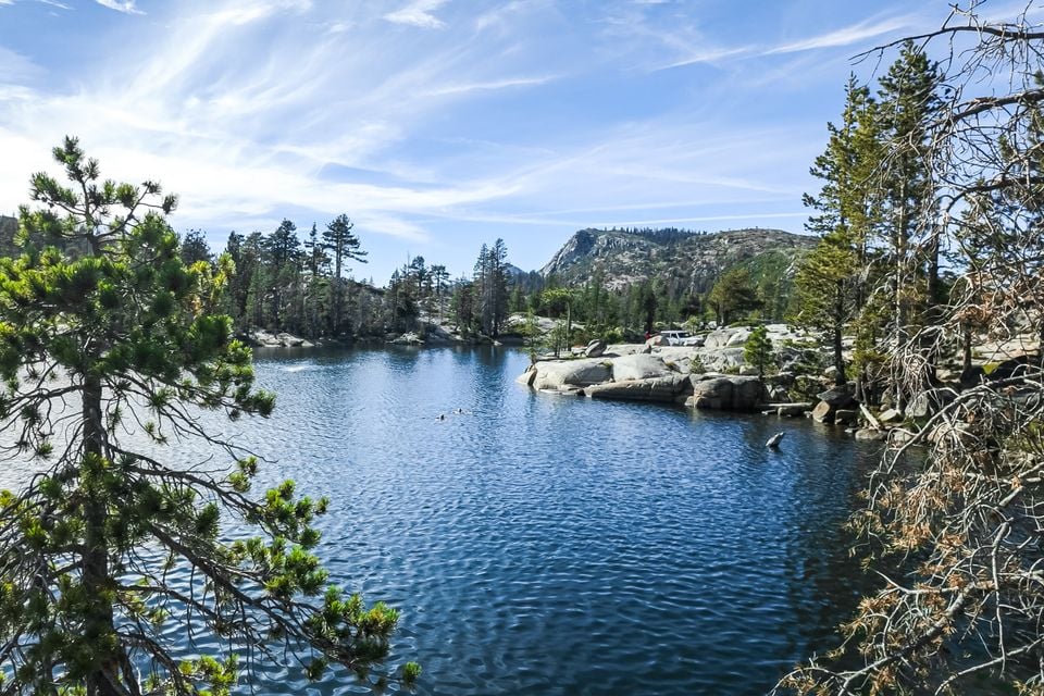An alpine lake surrounded by pine trees and granite boulders, with swimmers in the water and a mountain peak under a blue sky