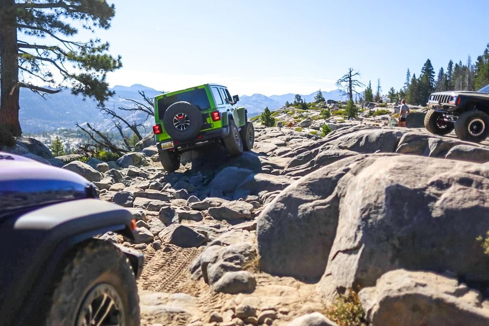 A green Jeep Wrangler 392 climbing a rocky section of the Rubicon Trail, seen from the rear, with other off-road vehicles and mountains nearby