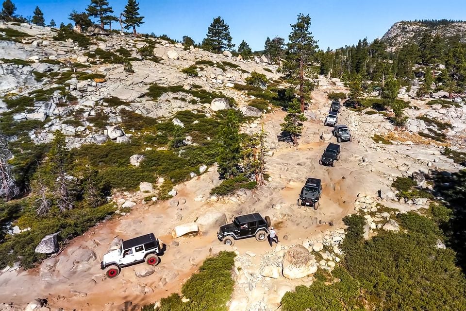 A convoy of Jeep Wrangler 4x4s crawling over granite rocks on the Rubicon Trail, seen from above amid pine trees