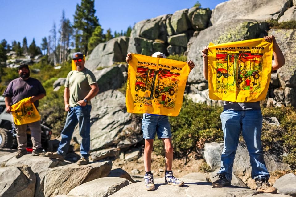 People standing on granite rocks holding up yellow Rubicon Trail 2026 bandanas, with a partly visible off-road vehicle in the background