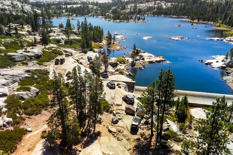 Aerial view of a convoy of Jeep Wrangler 392 off-roaders climbing granite slabs on the Rubicon Trail beside an alpine lake and pine forest