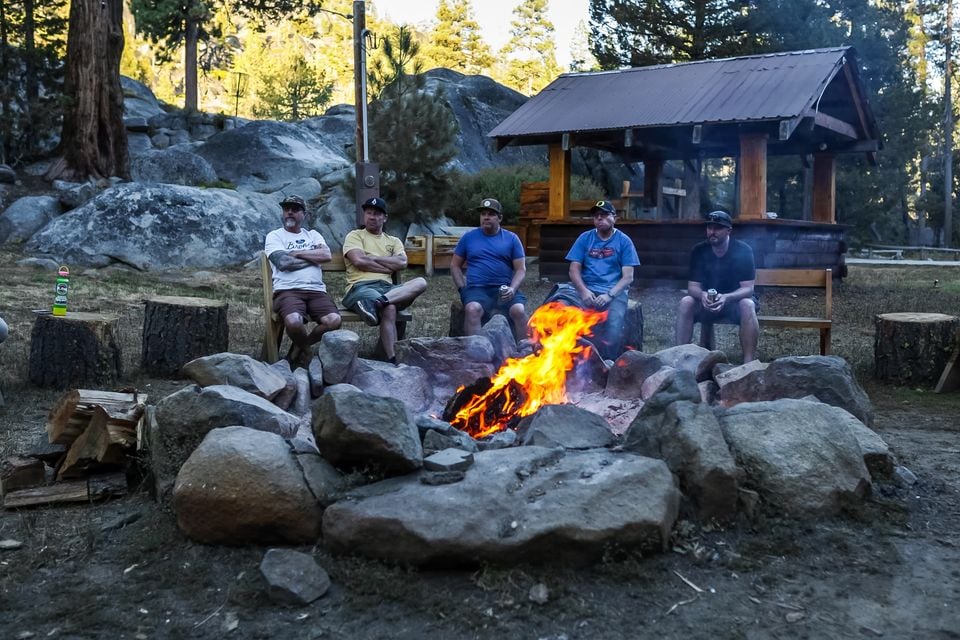 Five men in caps sit around a campfire in a stone fire pit at a forest campsite during the Jeep Wrangler 392 Rubicon Trail trip
