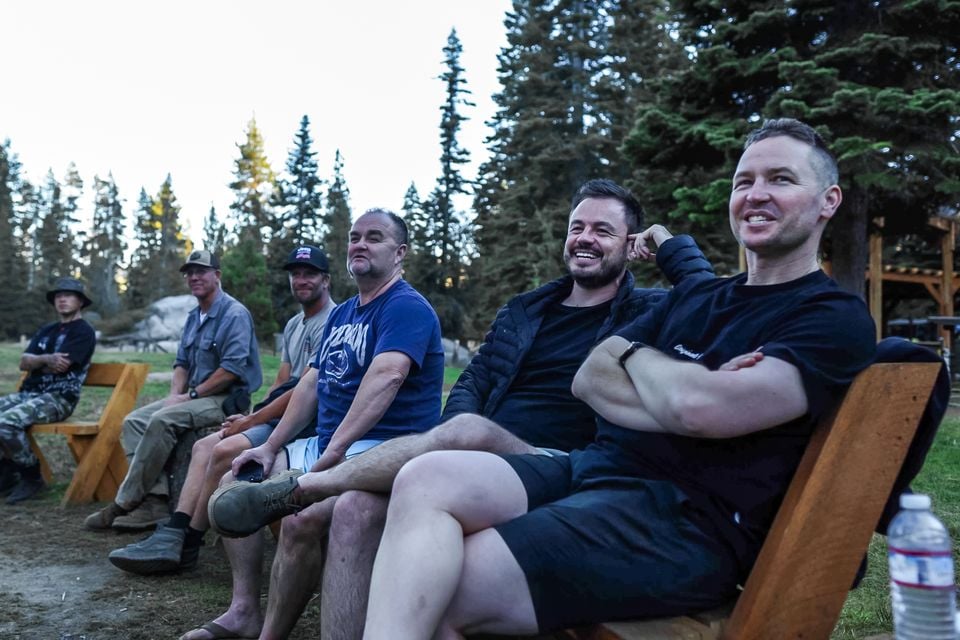 A group of men sitting on wooden benches at a forest campsite, smiling during a break on the Jeep Wrangler 392 Rubicon Trail trip