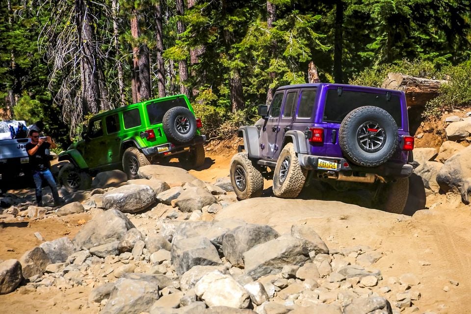 Purple and green Jeep Wrangler 392 four-door SUVs seen from the rear climbing over rocks on the Rubicon Trail in a pine forest