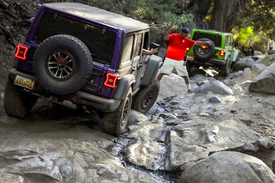 A purple Jeep Wrangler four-door seen from the rear crawling over rocks on the Rubicon Trail as a spotter guides it, with a green Wrangler ahead