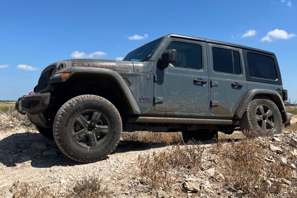 A mud-covered Jeep Wrangler Unlimited Rubicon SUV navigating rocky off-road terrain, seen from the front-side against a blue sky.