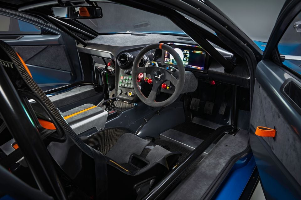 Interior of a race car showing a flat-bottomed steering wheel with button controls, carbon fibre trim, a digital display dashboard, pedal box, and roll cage structure