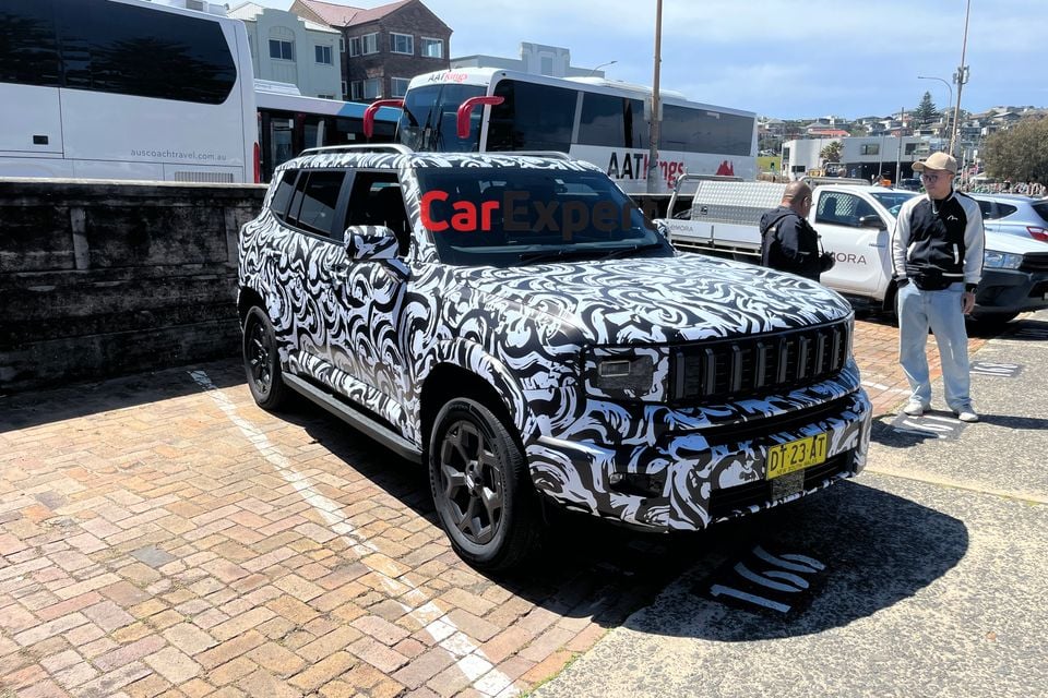 A camouflaged Jetour T1 SUV prototype parked on a brick-paved car park in Sydney, seen from the front, with onlookers nearby
