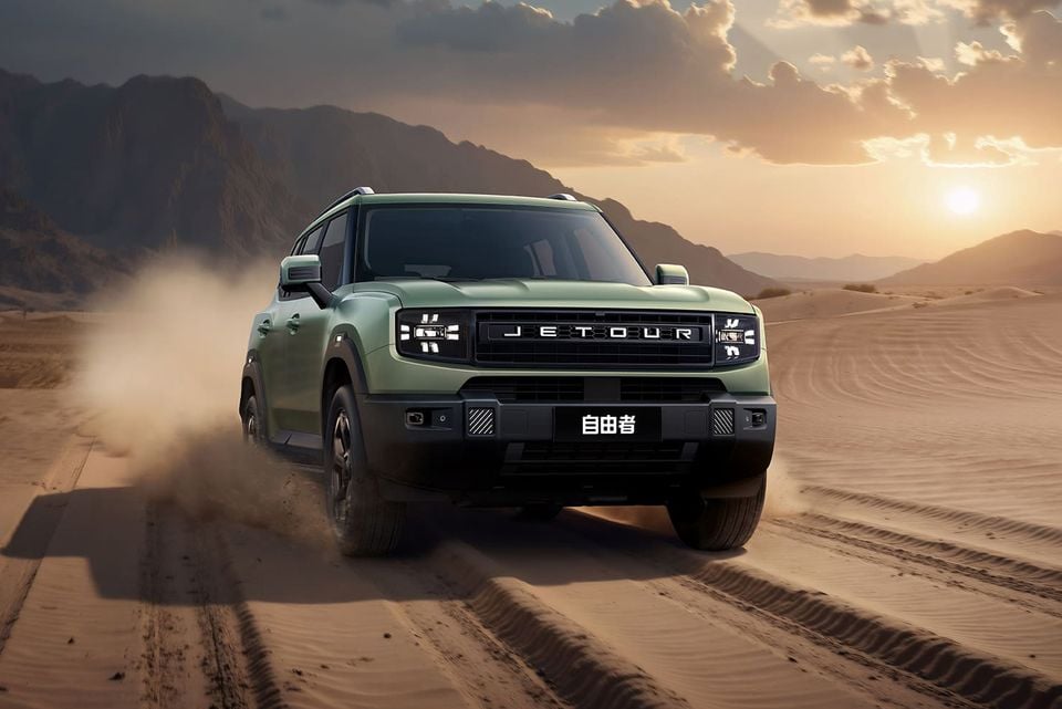 A green Jetour T1 SUV driving across desert sand dunes at sunset, seen from the front and kicking up dust