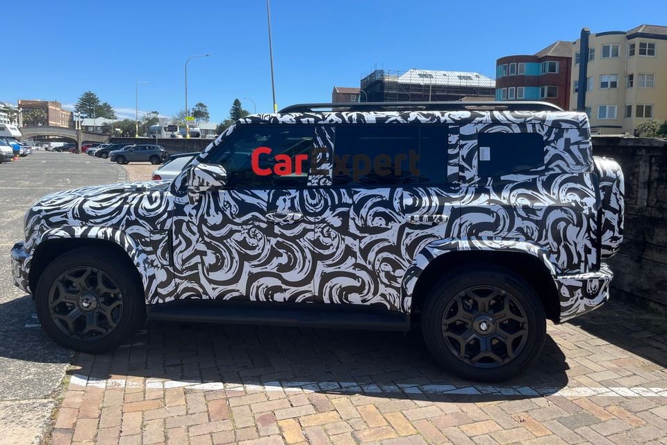 A camouflaged Jetour T2 SUV prototype seen side-on, parked on brick paving near a beachside car park in Australia