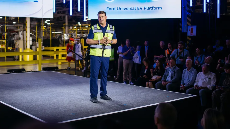 A man in a high-visibility vest speaks on stage at a Ford event, with a screen behind him displaying 'Ford Universal EV Platform' and an audience seated to the right inside a manufacturing facility.