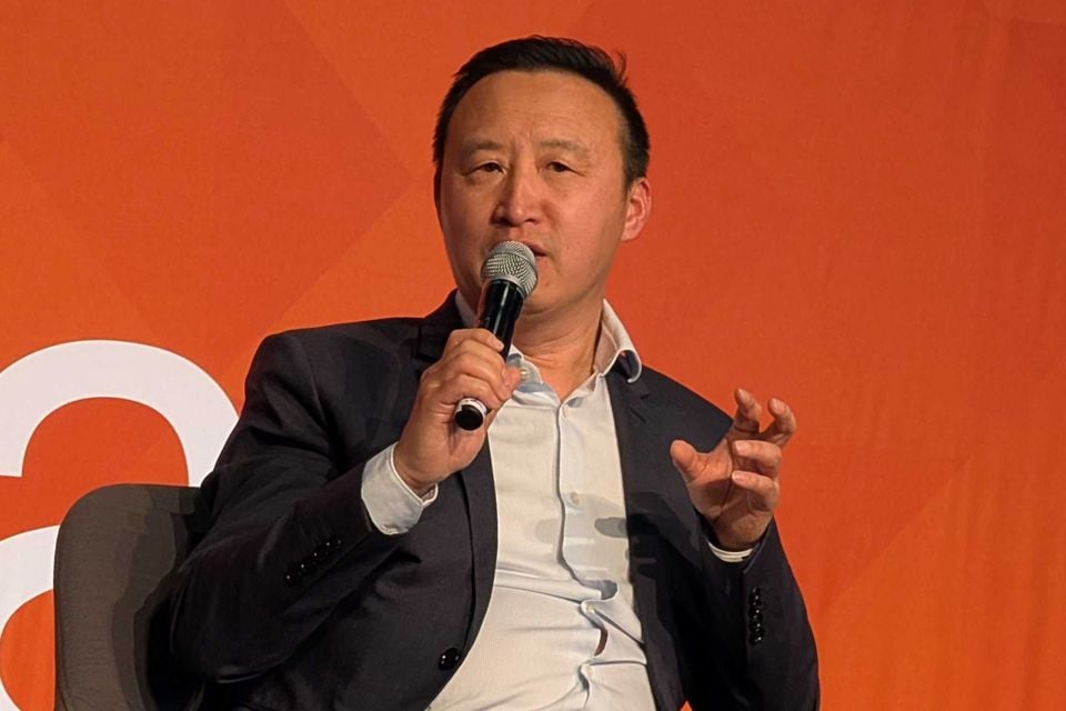 A man in a dark blazer speaks into a microphone at an event in front of an orange branded backdrop.