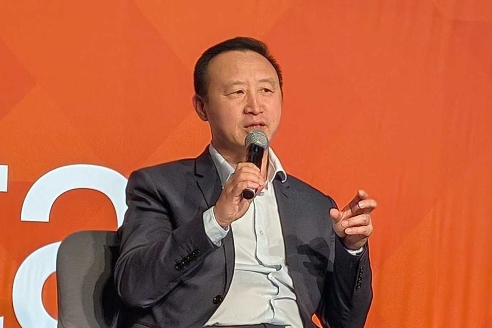 A man in a dark suit speaks into a microphone while seated on stage at an event, gesturing with his hand.
