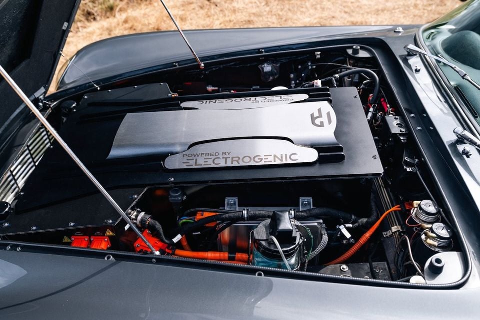 An electric motor conversion by Electrogenic fitted in the engine bay of a classic car, with orange high-voltage cables and a cover reading 'Powered by Electrogenic'